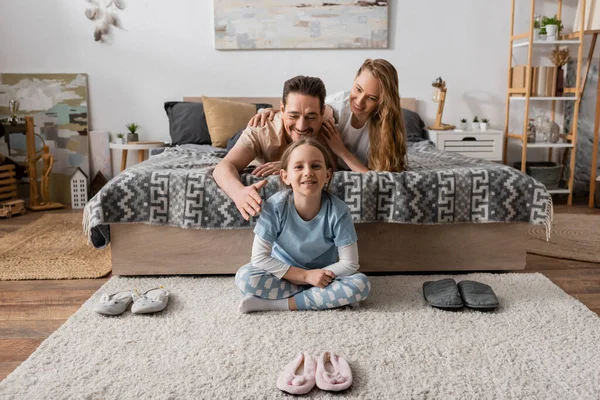 Положительный ребенок сидит на ковре вокруг тапочки рядом счастливые родители на кровати — стоковое фото