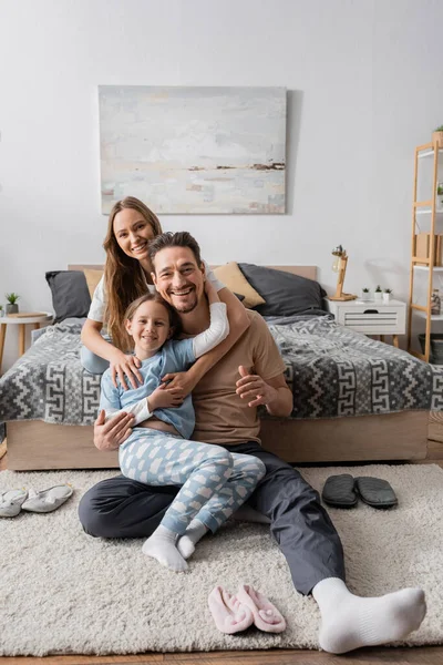Cheerful kid sitting with happy father on carpet around slippers near happy mother on bed — Stock Photo