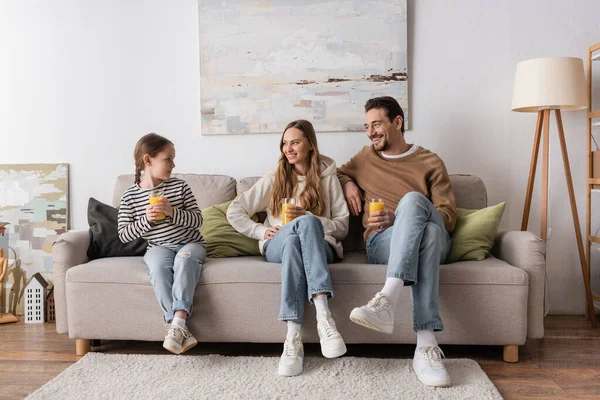 Full length of cheerful parents holding glasses of orange juice and looking at daughter while sitting on sofa — Photo de stock