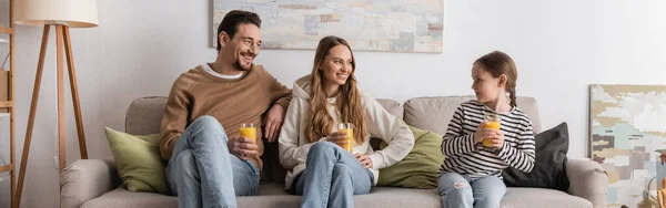 Cheerful parents holding glasses of orange juice and looking at daughter while sitting on sofa, banner — Photo de stock