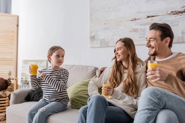 Cheerful parents holding glasses of orange juice and looking at daughter while sitting on couch — Photo de stock