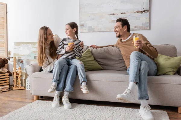 Full length of cheerful daughter sitting on laps of mother and holding glass of orange juice near happy father — Stockfoto