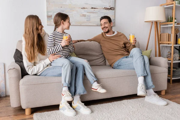 Full length of cheerful daughter sitting on laps of mother and holding glass of orange juice near positive father — Photo de stock