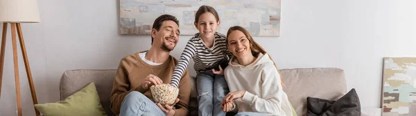 Happy kid with remote controller watching movie with cheerful parents, banner — Stock Photo