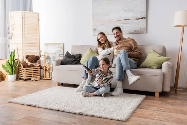 Full length of cheerful kid clicking channels near happy parents sitting with popcorn at home — Stock Photo