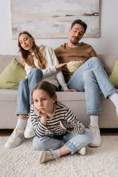 Full length of bored kid clicking channels near parents sitting with popcorn on blurred background — Stock Photo