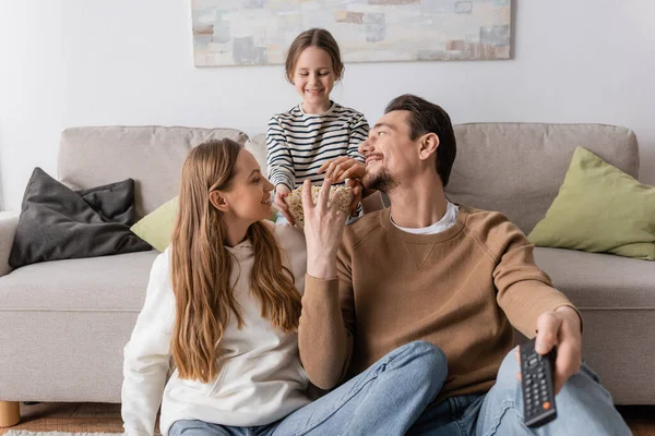 Happy man holding remote controller and reaching popcorn near wife and cheerful daughter on sofa — Stock Photo