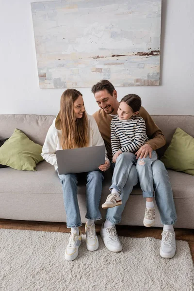 Full length of happy woman sitting with laptop near husband and daughter in living room — Fotografia de Stock