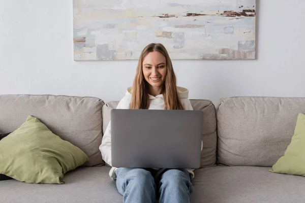 Cheerful woman in casual clothes using laptop while sitting on couch — Photo de stock