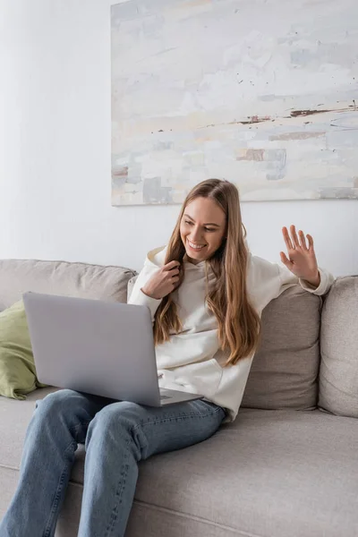 Positive freelancer in casual clothes waving hand while using laptop in living room — Foto stock