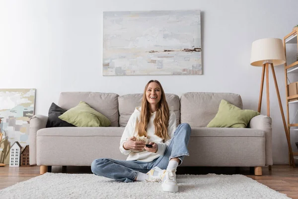 Full length of joyful woman holding remote controller while watching movie in living room — Stock Photo