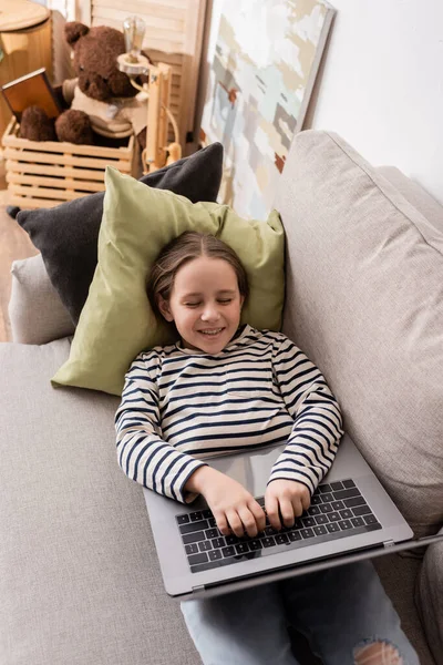 High angle view of smiling preteen girl in casual clothes using laptop while lying on couch — Stock Photo