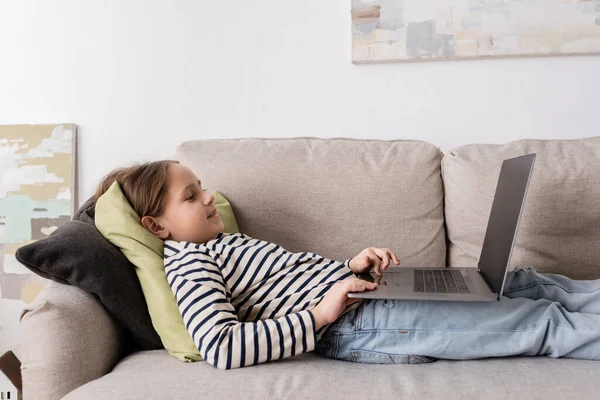 Side view of cheerful preteen girl in casual clothes using laptop while lying on couch — Stock Photo