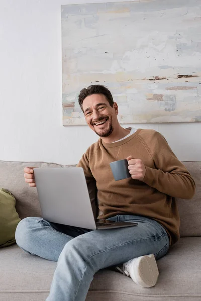 Joyful man using laptop while holding cup and sitting on sofa in living room - foto de stock