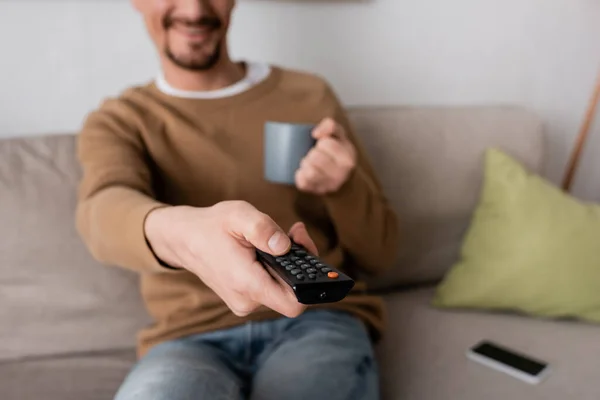 Cropped view of cheerful man holding remote controller and cup of coffee in living room — Stock Photo