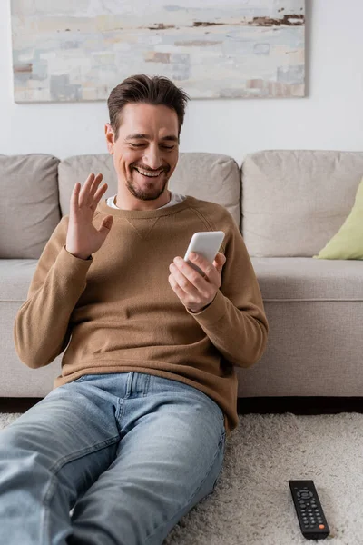 Happy man in beige jumper waving hand at smartphone while having video call at home — Foto stock