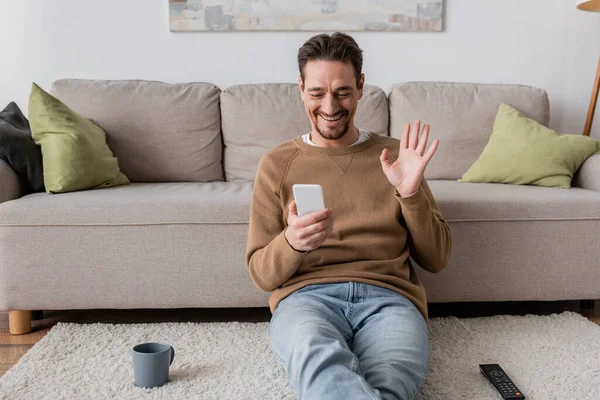 Cheerful man in beige jumper waving hand at smartphone while having video call at home — Foto stock