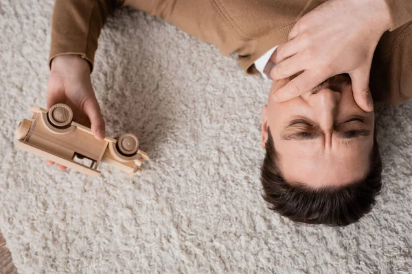 Top view of sad man covering mouth with hand lying on carpet near wooden toy vehicle in living room — Foto stock