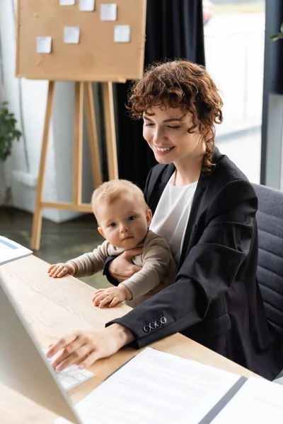Улыбающаяся деловая женщина, работающая за компьютером, сидя на рабочем месте с дочкой малыша — стоковое фото