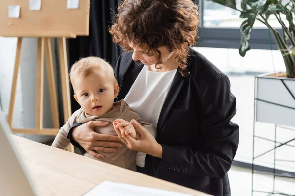 Businesswoman in black blazer holding hand of toddler child while sitting at workplace in office — Fotografia de Stock