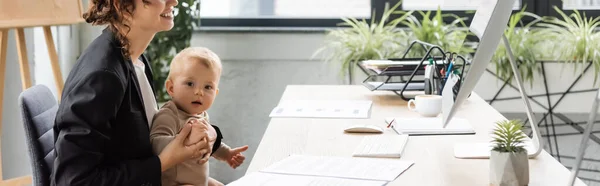 Toddler child looking at camera near smiling mother sitting at workplace near documents and computer monitor, banner — Stockfoto
