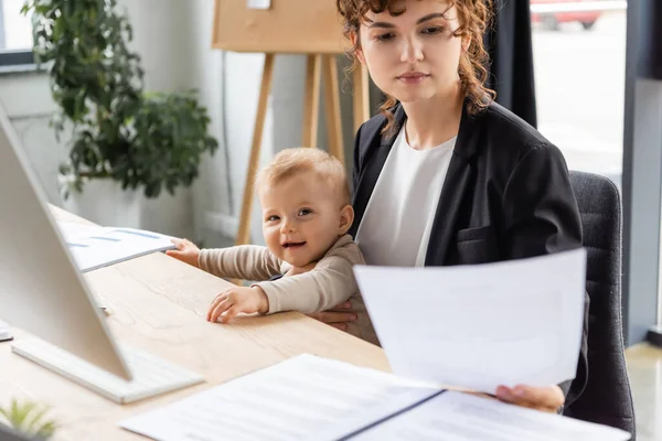 Businesswoman in black blazer holding document near cheerful toddler daughter at workplace in office — Stockfoto