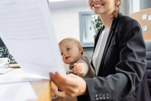 Joyful businesswoman holding blurred document near little child with outstretched hand in office — Stockfoto