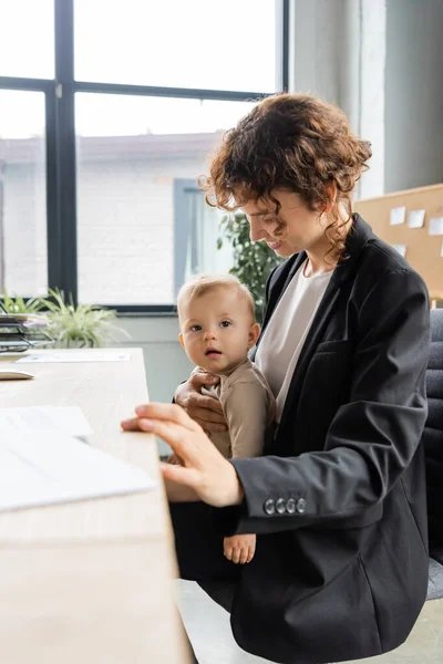 Улыбающаяся деловая женщина в черном блейзере, сидящая на рабочем месте с ребенком во время работы в офисе — стоковое фото