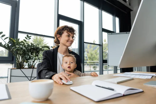 Smiling businesswoman working on computer near little daughter and blurred notebook on work desk — Stockfoto