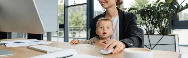Little girl looking at camera near smiling mom working on computer in office, banner — Stock Photo