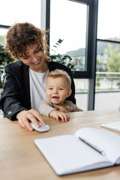 Smiling businesswoman holding computer mouse near little cheerful daughter and blank notebook on blurred foreground — Stockfoto