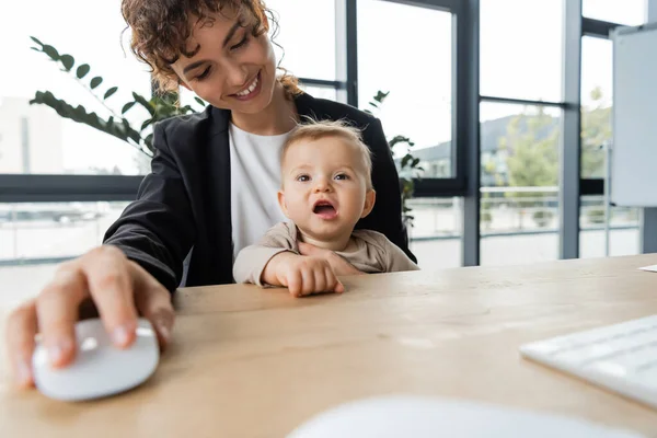 Cheerful businesswoman sitting at workplace with little child and holding blurred computer mouse — Stockfoto