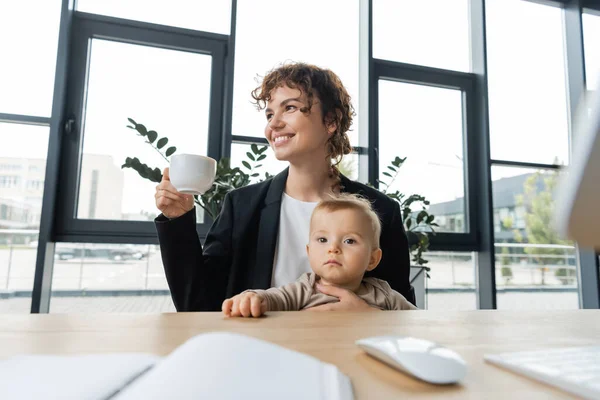 Happy businesswoman sitting with little daughter and coffee cup near blurred computer mouse and notebook on work desk — Stock Photo