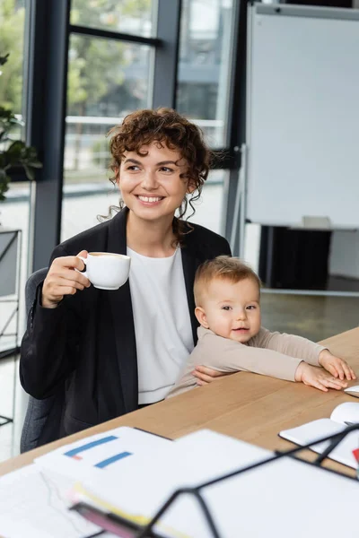 Улыбающаяся деловая женщина смотрит в сторону, сидя с ребенком малыша и чашки кофе на рабочем месте в офисе — стоковое фото