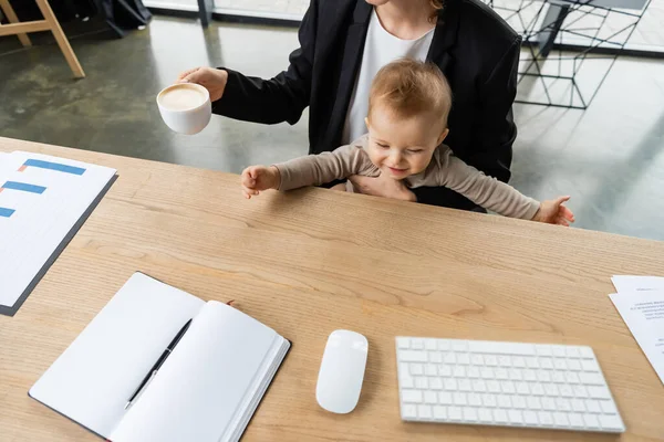 Высокий угол обзора веселого малыша рядом с матерью с чашкой кофе и компьютерной мышью с клавиатурой и пустой ноутбук на рабочем столе — стоковое фото