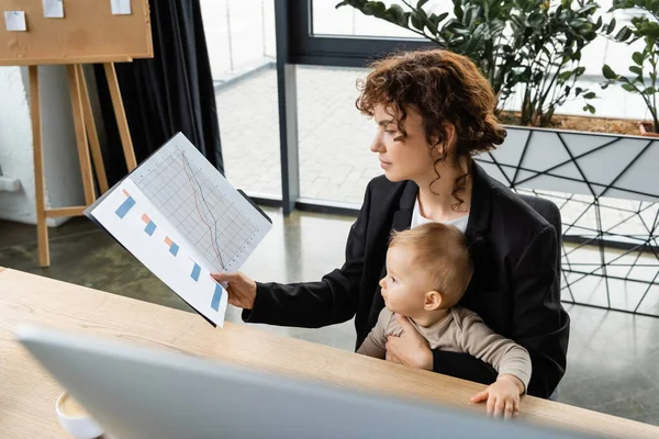 Деловая женщина в черном блейзере сидит на рабочем месте с маленькой дочерью и смотрит на папку с графиками — стоковое фото