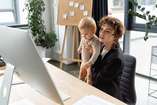 Бизнесвумен в черном пиджаке держит ребенка и смотрит на монитор компьютера в офисе — стоковое фото