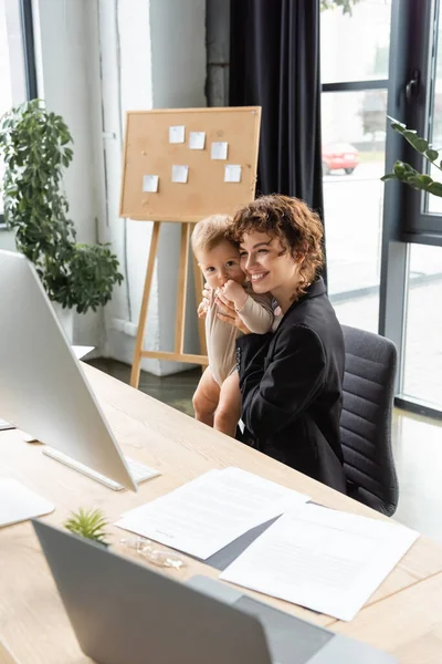 Pleased businesswoman holding toddler child while sitting near computers and documents in office — Stock Photo
