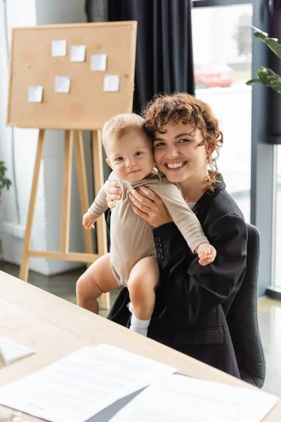 Cheerful businesswoman holding little daughter in romper while sitting at work desk near blurred documents — Stockfoto