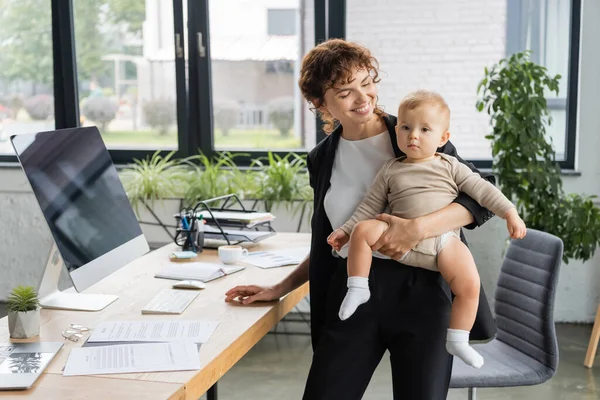 Smiling businesswoman holding toddler daughter in romper near work desk with computer monitor and documents — Stockfoto