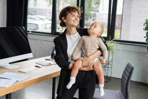 Счастливая деловая женщина смотрит в сторону, стоя с маленьким ребенком рядом с монитором компьютера с чистым экраном — стоковое фото