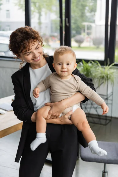 Joyful businesswoman in black suit holding toddler daughter in romper in office — Photo de stock