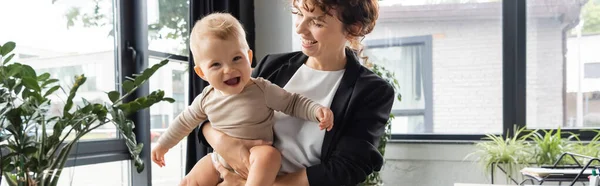 Smiling businesswoman holding cheerful toddler child in office, banner — Stockfoto