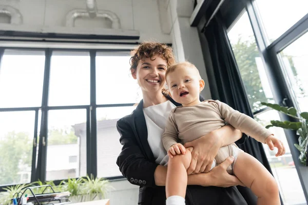 Joyful businesswoman looking at camera while holding baby girl in office — Stockfoto