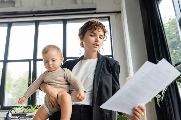 Office manager in black blazer looking at papers while holding toddler baby in office — Fotografia de Stock