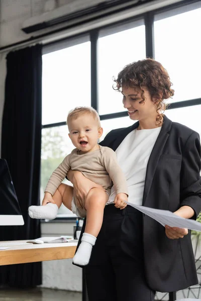 Pleased businesswoman in black suit holding papers and happy daughter in romper in office — Photo de stock