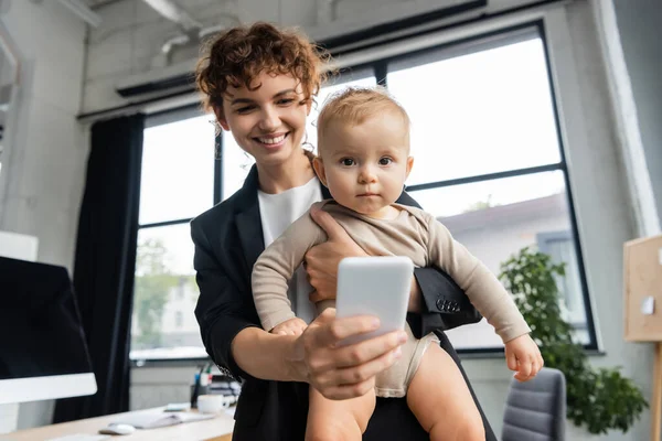 Smiling businesswoman with little child taking selfie on cellphone in office — Stock Photo