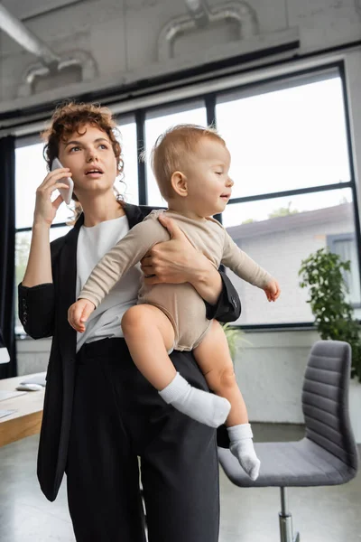 Деловая женщина в черном костюме разговаривает по мобильному телефону, держа малышку дочь в офисе — стоковое фото