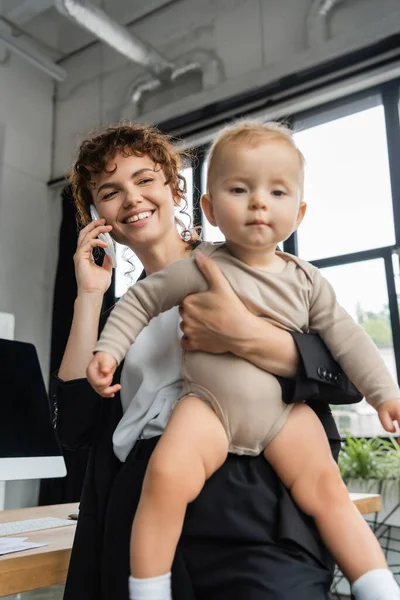 Positive businesswoman talking on smartphone and holding toddler child in office — Stock Photo