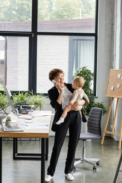 Smiling businesswoman in black suit holding little daughter and talking on mobile phone in modern office — Stockfoto